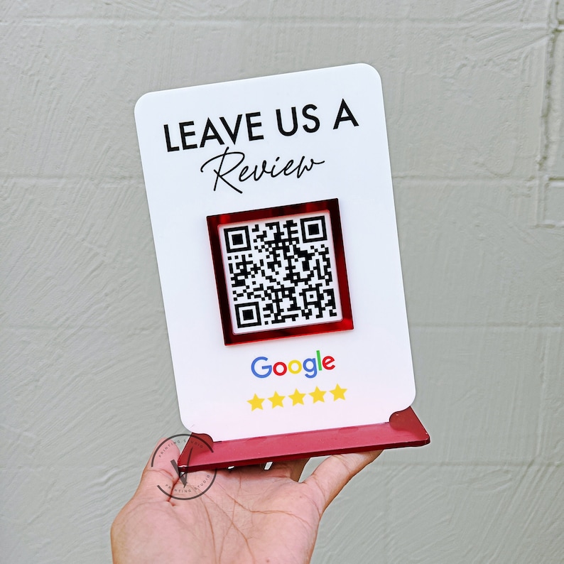 May include: White sign with the text "LEAVE US A Review" in black lettering. A red square with a black and white QR code is below the text. The text "Google" is below the QR code in black lettering. Five gold stars are below the text "Google". The sign is on a red stand.