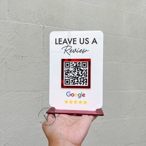 May include: White sign with the text "LEAVE US A Review" in black lettering. A red square with a black and white QR code is below the text. The text "Google" is below the QR code in black lettering. Five gold stars are below the text "Google".