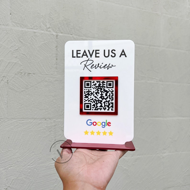 May include: White sign with the text "LEAVE US A Review" in black lettering. A red square with a black and white QR code is below the text. The text "Google" is below the QR code in black lettering. Five gold stars are below the text "Google".