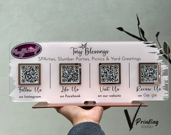 Op de afbeelding: Een wit acryl bord met de tekst "Tiny Blessings" en de woorden "SPArties, Slumber Parties, Picnics & Yard Greetings". Het bord heeft vier vierkante frames met QR-codes erin. Elk frame heeft een label eronder: "Follow Us on Instagram", "Like Us on Facebook", "Visit Us on our website", en "Review Us on Google". Het bord staat op een roségouden standaard met de tekst "Printing STUDIO" onderaan.