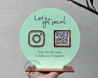 May include: A light green round sign with the text "Let's get social" in black. The sign has an Instagram icon and a QR code. The text below the icons reads "Scan the QR code to follow our Instagram".