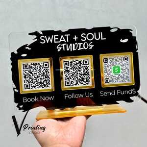 May include: A black acrylic sign with gold accents featuring the text "Sweat + Soul Studios" and three QR codes. The QR codes are labeled "Book Now", "Follow Us", and "Send Funds".