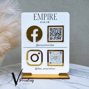 Könnte beinhalten: Ein weißes Schild mit goldenen Akzenten mit dem Text "EMPIRE HAIR" mit Social-Media-Symbolen und QR-Codes für Facebook und Instagram. Der Text "@empirehairdee" und "@dee_empirehair" befindet sich unter den Symbolen. Das Schild steht auf einem goldenen Ständer.