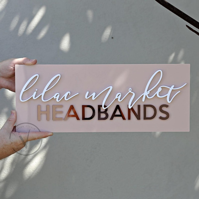 Rectangle acrylic business sign in blush pink acrylic backing installed on the wall, LILAC MARKET in 3D gloss white acrylic on top in cursive font, HEADBANDS under it in 3D rose gold mirror in SANS font