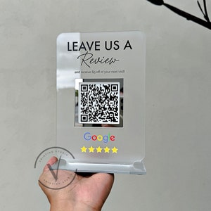May include: A clear acrylic sign with the text "LEAVE US A Review and receive £5 off of your next visit!"  A QR code is displayed below the text, followed by the word "Google" and five yellow stars.