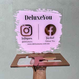 Puede incluir: Un letrero acrílico transparente con fondo rosa y base de oro rosa. El letrero presenta el texto "DeluxeYou" e iconos de redes sociales para Instagram y Facebook, junto con información del sitio web.