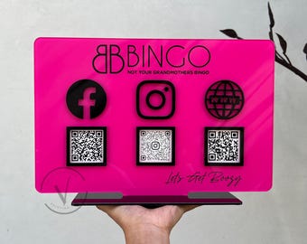Op de afbeelding: Een felroze acrylbord met de tekst "BB BINGO" en "NOT YOUR GRANDMOTHERS BINGO". Het bevat zwarte social media-iconen en QR-codes. De zin "Let's Get Boozy" staat onderaan.
