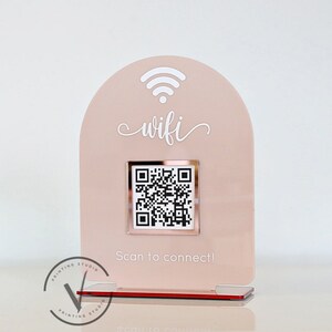 May include: A pink acrylic sign with a white wifi symbol and the word "wifi" in cursive. A rose gold square with a QR code is in the centre of the sign. The text "Scan to connect!" is below the QR code.