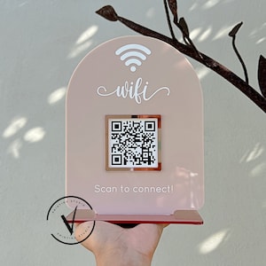 Op de afbeelding: Een roze acryl bord met een afgeronde bovenkant en een wit wifi-symbool. Het bord zegt "wifi" in witte cursieve letters. Een vierkant, zilveren frame met een zwart-witte QR-code bevindt zich in het midden van het bord. De tekst "Scan to connect!" staat onder de QR-code.