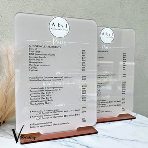 May include: Two white acrylic signs with black text listing prices for anti-wrinkle and dermal filler treatments. The signs are on a wooden stand with a rose gold finish.