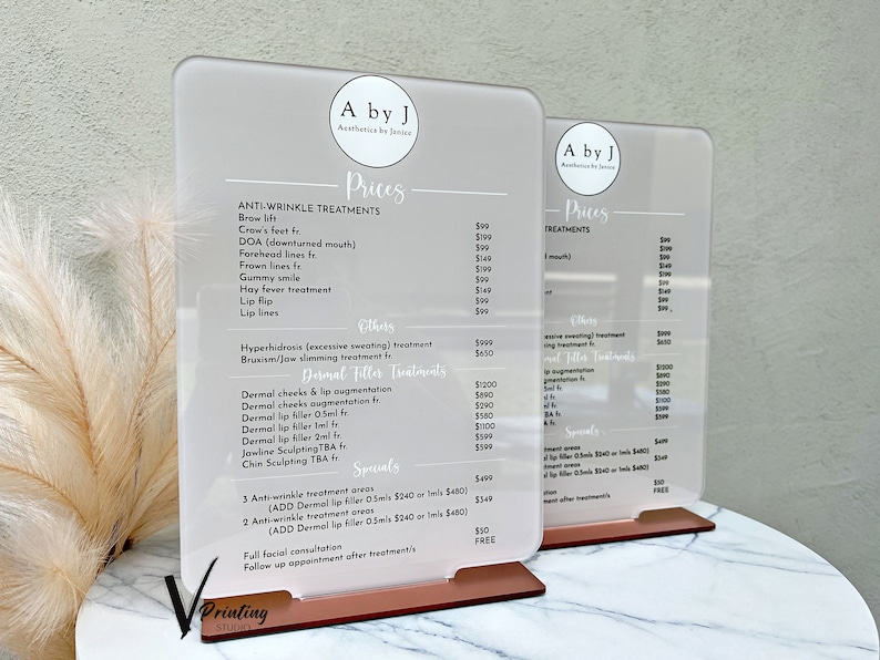 May include: Two white acrylic signs with black text listing prices for anti-wrinkle and dermal filler treatments. The signs are on a wooden stand with a rose gold finish.
