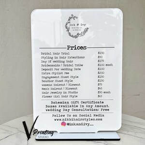 May include: A white sign with black text listing prices for hair styling services. The sign says "Ink & Ivy" and lists prices for services such as bridal hair trial, styling in hair extensions, day of wedding hair, bridesmaids, bridal tribe, deposit for wedding date, extra stylist fee, engagement shoot style, boudoir shoot style, women's haircut and blowout, men's haircut and blowout, hair jewelry in studio, and flower girl hair style.