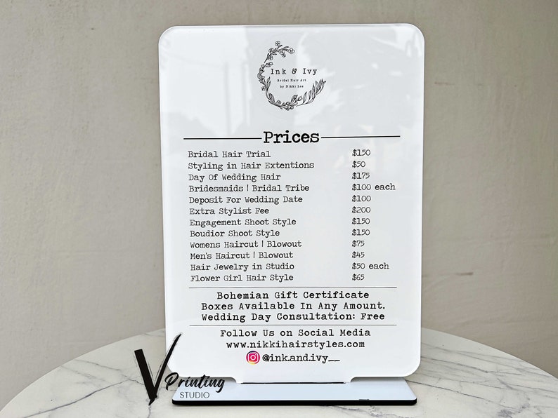 May include: A white sign with black text listing prices for hair styling services. The sign says "Ink & Ivy" and lists prices for services such as bridal hair trial, styling in hair extensions, day of wedding hair, bridesmaids, bridal tribe, deposit for wedding date, extra stylist fee, engagement shoot style, boudoir shoot style, women's haircut and blowout, men's haircut and blowout, hair jewelry in studio, and flower girl hair style.