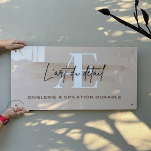 Rectangle acrylic business sign in blush pink acrylic backing installed on the wall, AE initial printed in white with brown splash, L art du detail across it in 3D black acrylic cursive font, business tagline in 3D taupe brown, 4 mounting holes