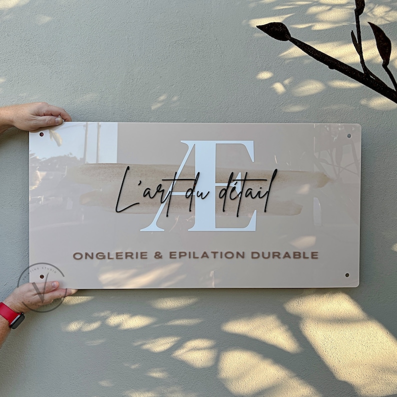 Rectangle acrylic business sign in blush pink acrylic backing installed on the wall, AE initial printed in white with brown splash, L art du detail across it in 3D black acrylic cursive font, business tagline in 3D taupe brown, 4 mounting holes