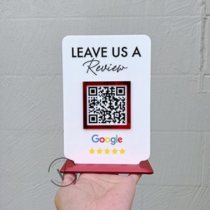 May include: White sign with red trim, reading "Leave Us A Review" in black text with a cursive "Review". A red square with a black and white QR code is below the text. Below the QR code is the word "Google" in black text and five gold stars.