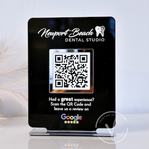 May include: Black acrylic sign with a white logo that says "Newport Beach Dental Studio" and a white tooth graphic. The sign also has a QR code and the text "Had a great experience? Scan the QR Code and leave us a review on Google" with five stars.