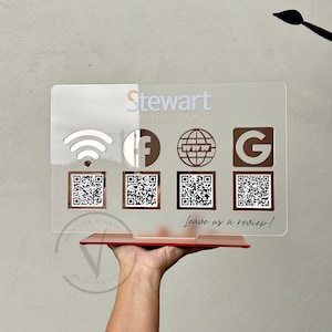 May include: A clear acrylic sign with the text "Stewart Family Law" in white lettering. The sign features icons for Wi-Fi, Facebook, a website, and Google. There are four QR codes below the icons. The sign has a red base and the text "Leave us a review!" is written in white.