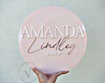 May include: A round, light pink sign with the name "Amanda Lindley Hair" in white and rose gold lettering.