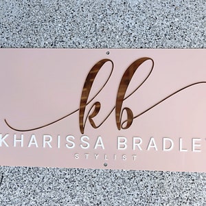 Custom Rectangle acrylic business sign in blush pink acrylic backing, initial KB in cursive font on top in 3D rose gold mirror, followed by KHARISSA BRADLEY under it in 3D gloss white acrylic in sans font, STYLIST on the bottom in sans font in white