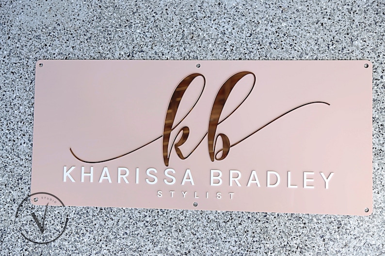 Custom Rectangle acrylic business sign in blush pink acrylic backing, initial KB in cursive font on top in 3D rose gold mirror, followed by KHARISSA BRADLEY under it in 3D gloss white acrylic in sans font, STYLIST on the bottom in sans font in white