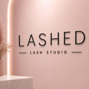 May include: A wall sign featuring the black, three-dimensional lettering LASHED LASH STUDIO in a clean, sans-serif font. The text is centred on a smooth, light pink wall, flanked by horizontal lines, with a vertical light strip to the left.