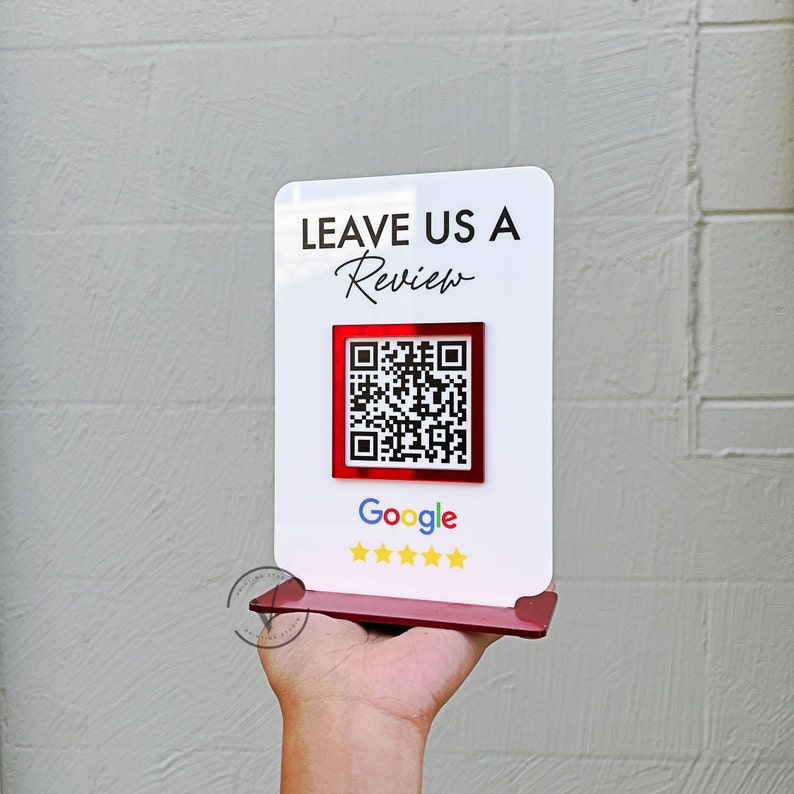 May include: White sign with the text "LEAVE US A Review" in black. A red square with a black QR code is below the text. Below the QR code is the word "Google" in black and five white stars.