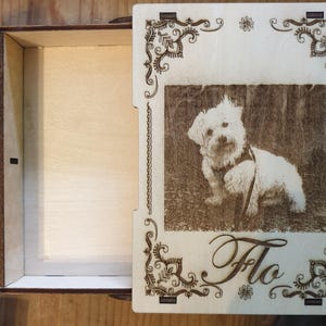 Könnte beinhalten: Eine Holzkiste mit einem Scharnierdeckel. Der Deckel ist mit einem dekorativen Muster und einem Foto eines weißen Hundes verziert, unter dem der Name "Flo" eingraviert ist.