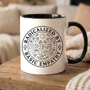 May include: A white ceramic mug with a black interior and handle. The mug features a circular design with the words "RADICALIZED BY BASIC EMPATHY" and various small illustrations in black.