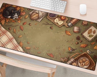 May include: A rectangular desk mat featuring an autumn picnic scene with a black and white cat, fallen leaves, a loaf of bread, fruit, a thermos, a mug, a book, a chipmunk, a bird, and a basket of cookies on a green grassy background.