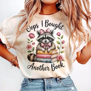 Könnte beinhalten: Cremefarbenes T-Shirt mit einem Waschbären, der eine rosa Schleife und Fliege trägt und auf einem Bücherstapel sitzt. Der Text "Oops I Bought Another Book" steht über und unter der Waschbär-Illustration, mit rosa Blumen auf beiden Seiten.