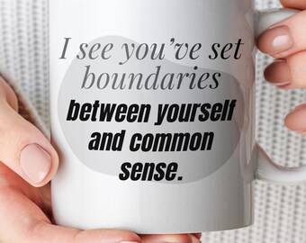 May include: A white ceramic mug featuring a gray organic-shaped background graphic with the black text: I see you've set boundaries between yourself and common sense. The mug has a standard handle and a smooth, glossy finish on the exterior surface.