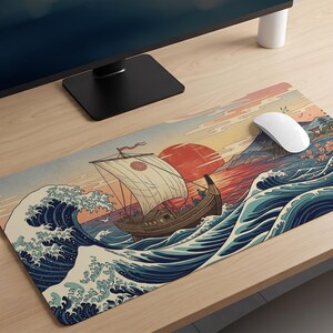 May include: A rectangular desk mat featuring a printed illustration of a wooden sailing ship navigating large, stylized blue ocean waves. The scene includes a large red sun in the background, a pale sky, and distant mountains. A white computer mouse sits on top.