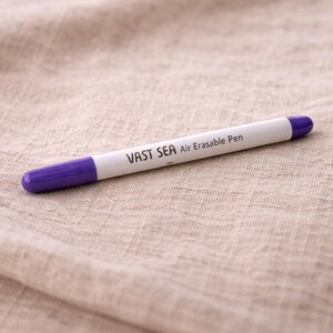 Pode incluir: Uma caneta apagável a ar branca com pontas roxas. A caneta tem o texto "VAST SEA Air Erasable Pen" impresso. Está sobre uma superfície de tecido bege.