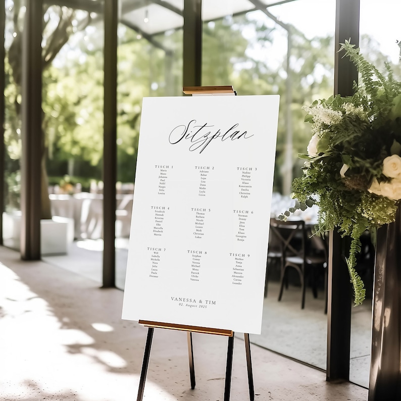 May include: A white seating chart with black text on an easel. The chart is titled "Sitzplan" and lists tables with names of guests. The chart is for Vanessa & Tim's wedding on August 2, 2023.