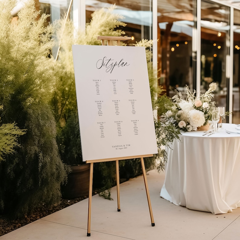 May include: A white seating chart on an easel with the word "Sitzplan" written in cursive at the top. The chart lists tables with names for each table. The chart is labeled "Vanessa & Tim" at the bottom.