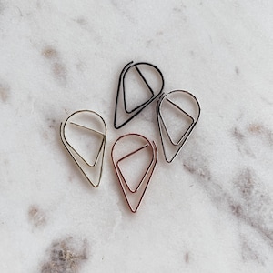 May include: Four metal paper clips in different colors: gold, silver, rose gold, and black. The clips are shaped like teardrops with a triangle inside.