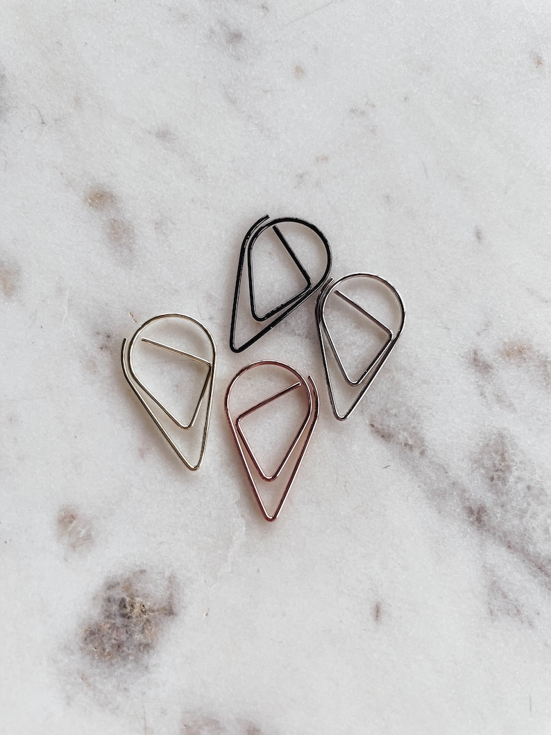 May include: Four metal paper clips in different colors: gold, silver, rose gold, and black. The clips are shaped like teardrops with a triangle inside.