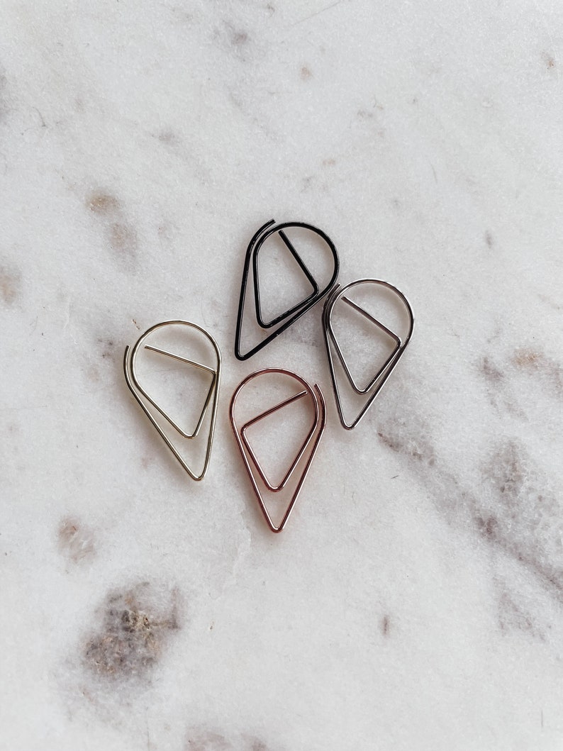 May include: Four metal paper clips in different colors: gold, silver, rose gold, and black. The clips are shaped like teardrops with a triangle inside.