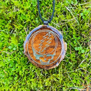 May include: A wooden pendant necklace featuring a Steal Your Face design. The pendant is a circular wood slice with a brown finish and a silver-colored graphic. The necklace has a black cord and is set against a green moss background.