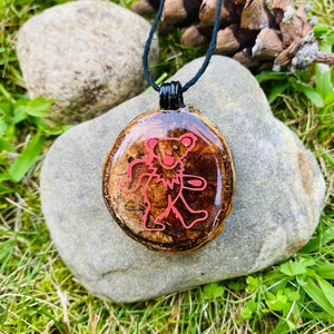 Puede incluir: Un collar con un colgante de madera rojo y marrón con un gráfico de oso Grateful Dead rojo. El collar está en un cordón negro.