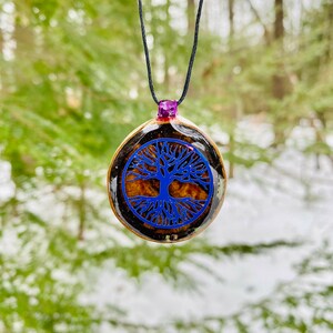 Puede incluir: Un colgante de árbol de la vida azul en un collar de cordón negro. El colgante está hecho de madera y resina y tiene un acabado transparente y brillante.
