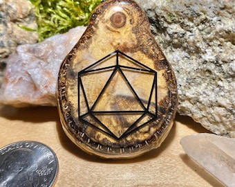 May include: A wooden pendant featuring a black resin d20 die design. The pendant is teardrop shaped and has a natural wood grain pattern. It is suspended from a black cord.