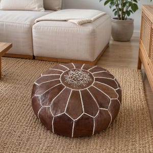 May include: A round, dark brown leather pouf featuring a flat top panel with intricate white embroidered geometric patterns. The sides are constructed from segmented leather panels joined by white stitching, creating a distinct floral-like shape.