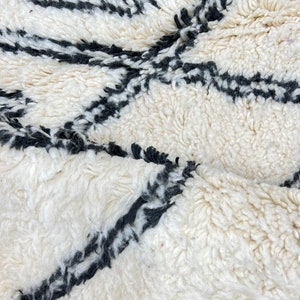 May include: White and black Moroccan rug with a geometric pattern. The rug is made of wool and has a soft, fluffy texture.