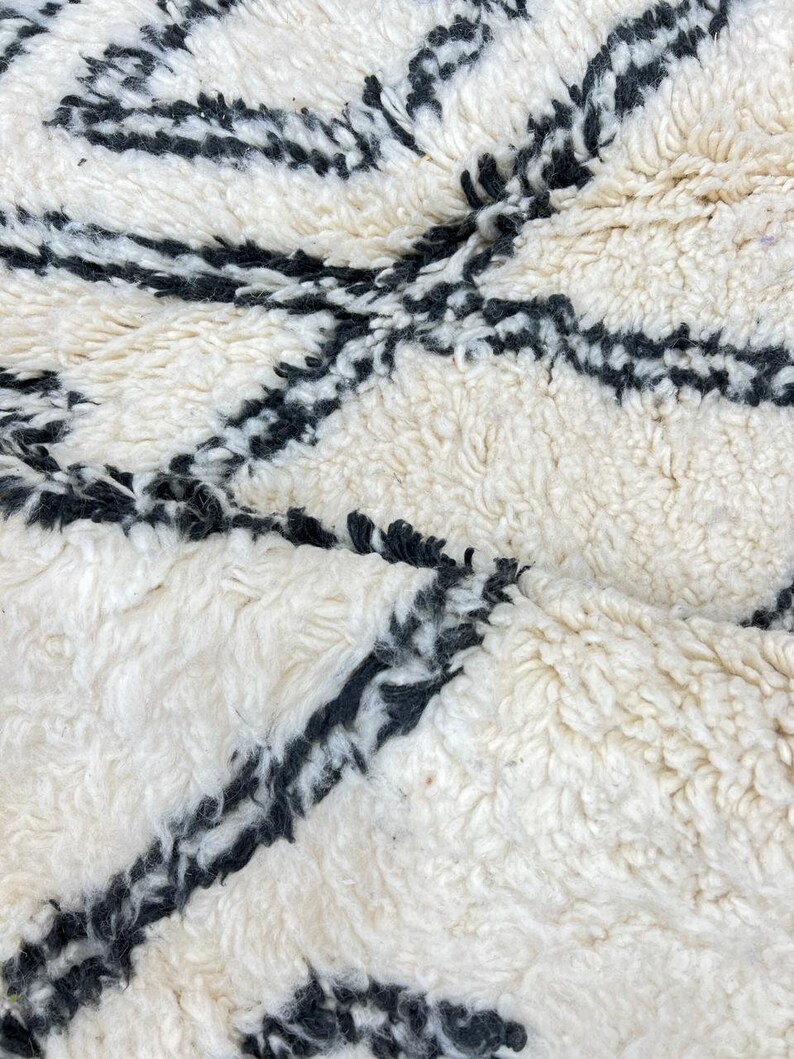 May include: White and black Moroccan rug with a geometric pattern. The rug is made of wool and has a soft, fluffy texture.
