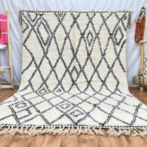 May include: A white and black Moroccan rug with a diamond pattern. The rug has a fringe border and is laid out on a wooden floor.