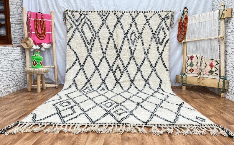 May include: A white and black Moroccan rug with a diamond pattern. The rug has a fringe border and is laid out on a wooden floor.