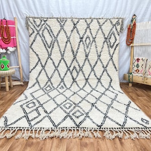 May include: A white and black Moroccan rug with a diamond pattern. The rug has a fringe border and is made of wool.