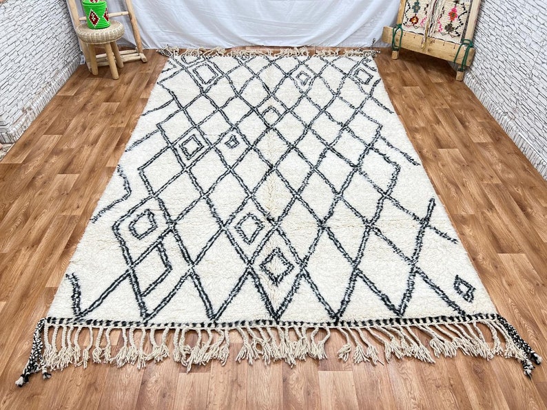 May include: A white and black Moroccan rug with a diamond pattern and fringe. The rug is made of wool and has a soft, fluffy texture.